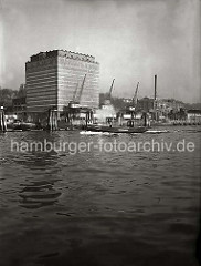 Das UNION KÜHLHAUS in Hamburg-Neumühlen; vor dem 1926 errichteten Gebäude stehen Kräne am Kai.  Eine Barkasse der Hafenpolizei fährt entlang der Duckdalben, die im Strom stehen.
