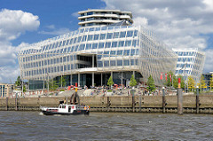 Elbseite vom Unileverhaus an der Norderelbe in der Hafencity Hamburgs - Touristen sitzen auf der Terrasse am Wasser in der Sonne, ein Sportboot fährt elbabwärts.