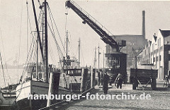 der Kai im Altonaer Hafen ist mit Kopfstein gepflastert - ein Kran holt die Ladung aus dem Laderaum des Frachtschiffs und lädt sie auf eine Anhänger der an Land steht; rechts Lagerhäuser. einige Hafenarbeiter sehen der Arbeit zu. (ca. 1939)