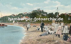 Badestrand am Elbufer von Blankenese ca. 1900; Mädchen in Kleidern stehen im Sand, andere bauen eine Sandburg. Eine Mutter liegt mit Kleid im Sand, dahinter ein Mann mit Anzug. Im Hintergrund die Häuser von Blankenese und der Süllberg.