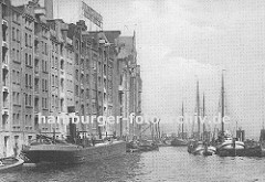 Blick auf die historische Hafenarchitektur der Speicher und Lagergebäude im Hafen von Altona; ein Motor-Binnenschiff wird mit Säcken beladen, die über die Winde am Dach des Lagerhauses auf das Schiff transportiert wird. (ca. 1925)