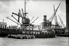 Ein Kaikran hievt Gefrierware auf das vor dem Neumühlener Kühlhaus liegende Frachtschiff. Auf den Pflastersteinen des Hafenkais liegen Metallfässer, ein Teil ist in die bereit stehenden offenen Güterwagen verladen. (ca. 1938)