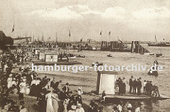 historisches Bild vom Strand an der Elbe bei Altona, Oevelgoenne. Frauen mit langen Kleidern und Hüten und Männer in Anzügen flanieren am Strand oder warten auf ein Ruderboot, das an den Verleihstationen am Elbstrand vermietet wird. Einige Ruderb