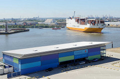 Empfangsgebäude / Abfertigungsgebäude vom Hamburger Cruise Center am Chicagokai an der Norderelbe - ein Frachter läuft in den Hamburger Hafen ein - er wird rückwärts von einem Schlepper in das Hafenbecken vom Hansahafen gezogen.