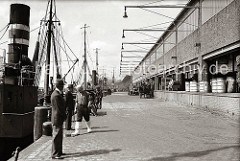 Rechts die 1937 neu gebaute Fischauktionshalle am Kai des Altonaer Fischereihafens -  Tonnen und Holzkisten zum Transport der Fische stehen auf der Laderampe des Gebäudes. Ein Pferdefuhrwerk wird beladen, dahinter ein Zugmaschine mit Anhänger, d