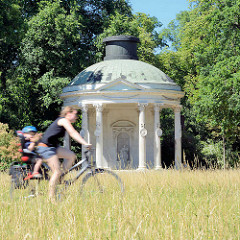 Fahrradfahrerin mit Kind, hohes Gras auf der Wiese vor dem Rundtempel im Potsdamer Park Sanssouci - der Freundschaftstempel ist ein kleiner Rundtempel, den der preußische König Friedrich II. zum Andenken an seine 1758 verstorbene Lieblingsschwester,