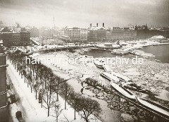 Altes Bild von Hamburg im Winter; Eis auf der Binnenalster, Schuten liegen an einem Steg - ein Schlepper unter Dampf. Im Hintergrund die verschneiten Geschäftshäuser am Jungfernstieg und die Kleine Alster mit der Schleusenbrücke.