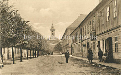 Blick durch die Meißler-Gasse in Theresienstadt zur Garnisonskirche - Strassenbäume; Geschäft für Kurzwaren und Stoffreste, Menschen vor der Eingangstür.