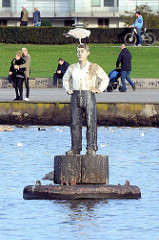 Bojenmann auf der Aussenalster vor dem Alsterufer bei Hamburg Hohenfelde - Holzskulptur, Bildhauer Stephan Balkenhol; eine Möwe sitzt auf dem Kopf, Spaziergänger am Ufer.