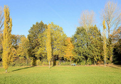 Bäume mit Herbstlaub und Parkbesucher auf einer Bank in Traunspark von Hamburg Rothenburgsort. Der Traunspark ist mit einer der ältesten Parks Hamburgs; im 18. Jhd. von Senator Friedrich Traun eingerichtet.