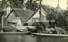 Spreewaldansicht früher - altes Foto von Holzhäusern und Schuppen mit Reet gedeckt; Spitze eines Heuschobers und Spreewaldkähne am Ufer.