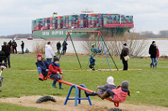 Kinderspielplatz in Grünendeich an der Elbe - Kinder auf der Schaukel und Wippe - im Hintergrund das auf einer Sandbank festgefahrene Containerschiff CSCL Indian Ocean.