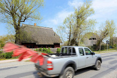 Reetdachhäuser an der Dorfstraße von Ahrenshoop - Pickup in Fahrt mit flatternden roten Fischereiflaggen.