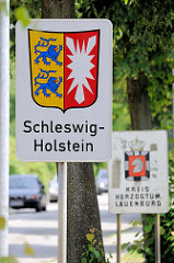 Schild Landesgrenze Schleswig-Holstein /  Wappen. Das Schleswig-Holsteiner Wappen vereint die Symbole der beiden ehemaligen Herzogtümer Schleswig und Holstein: Zwei rot bewehrte Löwen auf goldenem Grund und ein silbernes Nesselblatt auf rotem Grund.