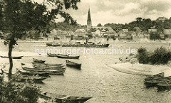 Historische Ansicht - Ruderboote / Fischerboote im natürlichen Hafen bei Hohnstorf an der Elbe, ein Binnenschiff fährt elbaufwärts. Altstadt von Lauenburg und Kirchturm der Maria Magdalenen Kirche.