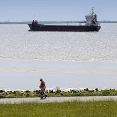 Blick vom Elbdeich bei Brunsbüttel - ein Tankschiff fährt Richtung Schleuse; eine Nordic Walkerin walkt am Ufer der Elbe.