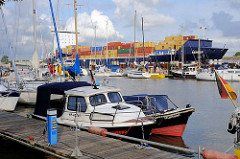 Sportboothafen / Yachthafen Brunsbütte - hinter der Schleuse im Nord-Ostsee-Kanal; Motorboote und Segelboote liegen am Steg, im Hintergrund fährt ein Containerfeeder in die Schleuse ein.