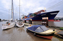 Sportboothafen / Yachthafen Brunsbütte - hinter der Schleuse im Nord-Ostsee-Kanal; Motorboot und Segelboote liegen am Steg, im Hintergrund fährt ein Containerfeeder in die Schleuse ein.