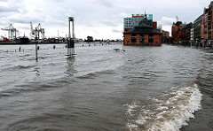 Das Hochwasser der Elbe hat die Kaimauer überspült und das Gelände des Hamburger Fischmarkts unter Wasser gesetzt. Auf der linken Bildmitte sind die Geländer der Kaianlage zu erkennen, dahinter die Wassertreppe zum Anleger, die teilweise unter Wa