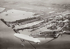 Alte Luftaufnahme des Hafens von Cuxhaven; ein Luftschiff fährt über dem Hafen. Im Hintergrund das Steubenhöft mit dem Amerikahafen und den Auswandererhallen am Kai. Davor die Einfahrt zum Alten Fischereihafen. Im Vordergrund die Seebäderbr