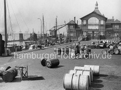 Historische Fischauktionshalle auf dem St. Pauli Fischmarkt in Hamburg.  Fässer liegen auf dem Platz - links eine Waage. Fischerboote haben am Kai festgemacht - im Hintergrund ein Kran und die Wassertreppe vom St. Pauli Fischmarkt.