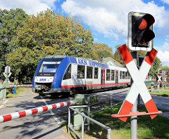 Bahnübergang bei der AKN Haltestelle Burgwedel in Hamburg Schnelsen. Die Schranken über Fußweg und Straße sind herunter gelassen, ein AKN Zug fährt Richtung Eidelstedt.