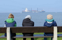 Elbufer - Blick auf die Elbe bei Otterndorf; Touristen sitzen auf einer Bank und blicken auf den Fluß - ein Containerschiff fährt in die Elbe ein.