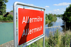 Rotes Stadtteilschild mit weisser Schrift, Allermöhe - plattdeutsch Allermeuh; im Hintergrund der Schleusengraben im Bezirk Hamburg Bergedorf.