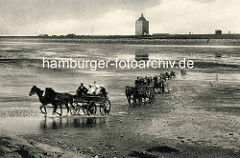 Historisches Foto von Wattwagen im Wasser - Personenwagen mit Touristen von Pferden gezogen auf den Weg von der Insel Neuwerk nach Duhnen / Cuxhaven. Im Hintergrund der Leuchtturm und Wohnhäuser hinter dem Deich.