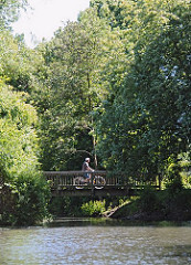 Ein Fahrradfahrer fährt über eine Holzbrücke entlang des Wanderweges am Ufer des Assmannkanals. Das Kanalufer ist mit Bäumen und Büschen dicht bewachsen.