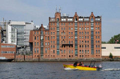 Ein gelbes Sportboot mit Aussenbordmotor fährt auf der Billwerder Bucht Richtung Sperrwerk. Am Ufer das historische Speichergebäude mit der roten Ziegelfassade.
