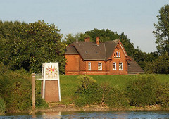 Eine Pegeluhr steht am Ufer der Norderelbe bei Kaltehofe, die Zeiger sind überklebt - die Anzeige funktioniert nicht mehr. Hinter dem Deich ragt eines der Verwaltungshäuser der ehem. Stadtwasserkunst.