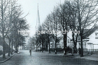 Marktplatz von Hamburg Barmbek, Straße mit Kopfsteinpflaster  und einem Radfahrer, links das Dach  eines Reetdachhauses - rechts Doppelhäuser.