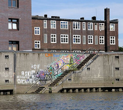 Im Billehafen führt eine Treppe hinunter zum Wasser des Hafenbeckens - die Wand ist mit buntem Graffiti bedeckt. Hinter der Kaimauer die dunkelroten Klinker eines der historische Gebäude am Billehafen.