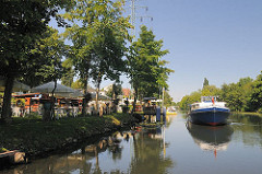 Biergarten am Ufer des Ernst-August-Kanals; Sonnenschirme und Tische stehem auf der Wiese am Wasser.