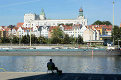 Ein Angler sitzt am Ufer der Oder - am Ufer liegt ein Binnenschiff,  dahinter eine Häuserzeile mit Hotel und das Stettiner Schloss mit den grünen Turmhelmen.
