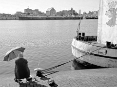 Ein Angler mit Sonnenschirm sitzt an der West-Oder im Hafengebiet von Stettin am Kai; am gegenüberliegenden Flussufer ein Binnenschiff und Hafenanlagen.