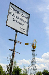 Hinweisschild Gose-Elbe, nicht schiffbares Gewässer - im Hintergrund  die Windpumpe " Die Hohe", eine 18 Meter hohe windbetriebene Wasserpumpe.