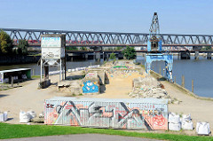 Blick zur Kaianlage mit Schüttgut am Billhafen in Hamburg Rothenburgsort; eine S-Bahn fährt über die Brücke am Oberhafenkanal.