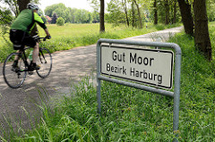 Ortsschild vom Hamburger Stadtteil Gut Moor, Bezirk Harburg - ein Fahrradfahrer auf einer Tour durch die Felder in Hamburgs Randbezirk.