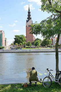 Blick über den Fluß Elbing zur Promenade und der katholischen St. Nikolaikirche - Baubeginn 13. Jahrhundert. Ein Angler sitzt am Flussufer, sein Fahrrad steht an einen Baum gelehnt.