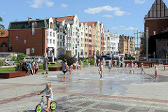 Kirchenvorplatz  mit Brunnen in Elbląg / Elbing - Kinder spielen in der Sonne - im Hintergrund Neubauten mit teilw. historisierenden Fassaden.