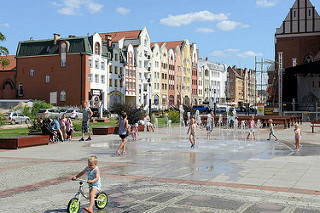 Kirchenvorplatz  mit Brunnen in Elbląg / Elbing - Kinder spielen in der Sonne - im Hintergrund Neubauten mit teilw. historisierenden Fassaden.