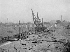Bau vom Baakenhafen in dem jetzigen Hamburger Stadtteil Hafencity - Bauarbeiter / Baumaschinen heben das Hafenbecken aus; lks. Masten von Schiffen in der Norderelbe, im Hintergrund die Gasanstalt auf dem Grasbrook.