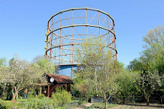 Gasometer in Münster, errichtet 1954 - stillgelegt 2005; die Glockenkonstruktion des Teleskopgasbehälters steht als technisches Denkmal unter Denkmalschutz. Im Vordergrund eine Kleingartenanlage mit Obstbäumen und Schrebergartenhaus.