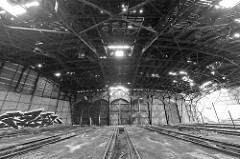 Einfahrten / Türen - teilweise zugemauert - eines Ringschuppens der Eisenbahn; Schienenanlage mit Grube für Arbeiten unter der Lokomotive. Das Dach des Lokschuppens ist stark verfallen.