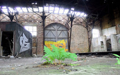 Türen - teilweise zugemauert - eines Ringschuppens der Eisenbahn; das Dach des Lokschuppens ist stark verfallen. Farn wächst aus dem Betonboden.
