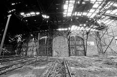 Türen - teilweise zugemauert - eines Ringschuppens der Eisenbahn; Schienenanlage mit Grube für Arbeiten unter der Lokomotive. Das Dach des Lokschuppens ist stark verfallen.