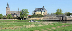Blick über die Maas auf die Burgruine De Keverberg und die Dorfkirche von Kessel; ein Sportboot fährt flussabwärts.