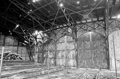 Einfahrten / Türen eines Ringschuppens der Eisenbahn; Schienenanlage mit Grube für Arbeiten unter der Lokomotive. Das Dach des Lokschuppens ist stark verfallen.